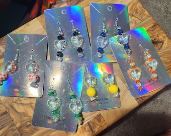 May include: Assortment of handmade earrings with various colored beads and clear heart-shaped charms. The earrings are displayed on cards with the text "Handmade with love".