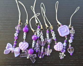 May include: A collection of purple and clear beaded charms with white cords. Charms include bows, hearts, butterflies, and stars. The word "BTS" is spelled out with white letter beads on several charms.