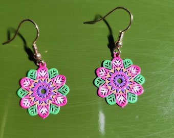 May include: A pair of colorful floral earrings. Each earring features a flower-shaped design with pink, green, purple, and yellow accents. The earrings have gold-colored ear wires and are set against a green background.