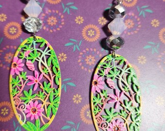 May include: A pair of oval-shaped earrings with a floral design. The earrings are made of metal and have a green, pink, and yellow color scheme. The earrings are attached to a silver chain with crystal beads.