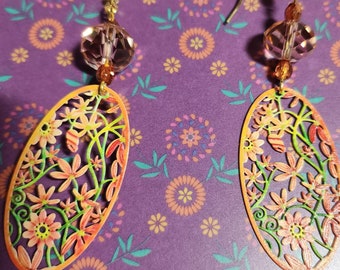 May include: A pair of dangle earrings with a floral design. The earrings are made of metal and have a gold finish. The floral design is cut out of the metal and painted in shades of orange, yellow, and green. The earrings are accented with a clear crystal bead.