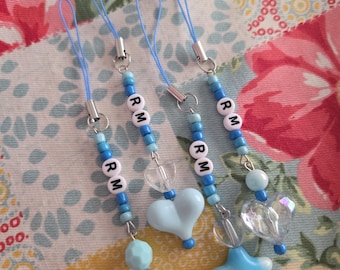 May include: Four blue beaded phone charms with the letters "RM". Each charm features a variety of blue beads, a heart, a star, and a clear bead. The charms have blue cords and silver-colored metal clasps.