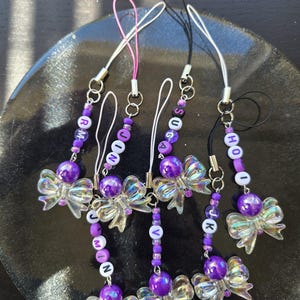 May include: A collection of six beaded phone charms. Each charm features a clear, iridescent bow, purple beads, and letter beads spelling out names. The charms have silver-colored hardware and colorful cords.