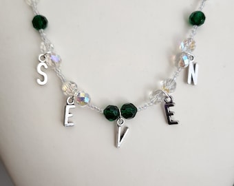 May include: A silver necklace with the word "SEVEN" spelled out in silver charms. The necklace is made with clear and green crystal beads.