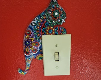 May include: A colorful mosaic cat figurine is mounted on a red wall next to a white light switch. The switch is in the "on" position.