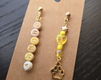May include: A pair of mismatched dangle earrings featuring gold-tone hardware. One earring has round beads spelling the name CHOMMY in yellow and white tones. The other earring features a small gold star, yellow beads, and a gold paw print charm.