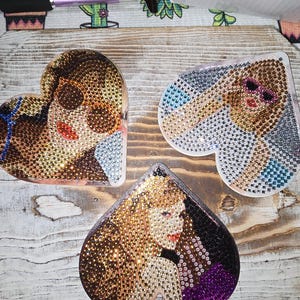 May include: Three heart-shaped images of women covered in rhinestones. Each image is a different colour and design. The images are on a wooden surface.