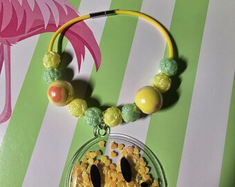 May include: A yellow circular necklace featuring a clear pendant filled with small yellow heart-shaped confetti and a black smiley face graphic. The necklace is accented with a series of round and textured beads in shades of yellow and light green.
