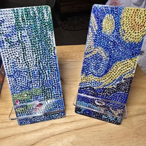 Puede incluir: Dos soportes para teléfono con una base de acrílico transparente y un diseño de mosaico colorido. Los diseños de mosaico son una escena de noche estrellada y un diseño abstracto verde y azul.
