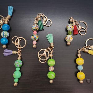 May include: A set of seven colorful wooden bead keychains with gold metal accents. Each keychain features a different pattern of colorful beads and a tassel.