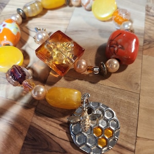 Peut inclure: Un bracelet en perles jaunes, orange et rouges avec un charme en verre transparent et un charme en argent avec un motif d'abeille et de nid d'abeille.