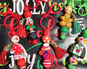 May include: Five Christmas-themed keychains with red, green, and white beads. Each keychain features a different charm, including a stocking, a Santa hat, a gingerbread man, a candy cane, and a sweater. The keychains are attached to silver key rings.