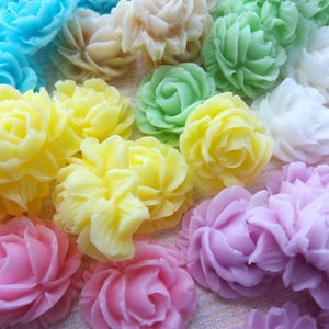 Può includere: Una collezione di saponi colorati a forma di fiore intagliato, nei toni del blu, giallo, rosa, verde e bianco. I saponi sono disposti vicini, mostrando i loro dettagliati disegni a petalo. Il tema generale è floreale e decorativo.