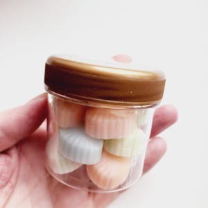 May include: A clear plastic jar with a brown lid filled with pastel-colored, shell-shaped candies. The candies are in shades of pink, blue, yellow, and orange. The jar is held in a person's hand.