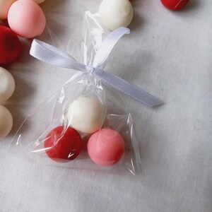 Puede incluir: Una bolsa de plástico transparente llena de tres caramelos redondos. Los caramelos son rojos, rosados y blancos. La bolsa está atada con una cinta blanca.