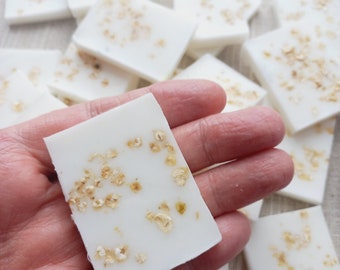 May include: A hand holds a white bar of soap with flecks of gold glitter. The soap is surrounded by other bars of soap with gold glitter.