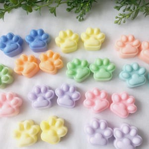 May include: A selection of pastel-coloured paw-shaped soaps in shades of blue, yellow, orange, green, pink, and purple. The soaps are arranged on a white surface, with green foliage in the background. These soaps are ideal for pet lovers.