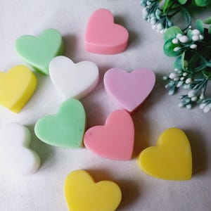 Può includere: Assortimento di saponi a forma di cuore in vari colori, tra cui rosa, verde, giallo e bianco. I saponi sono disposti su una superficie bianca.