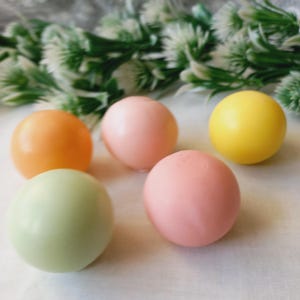Puede incluir: Cinco huevos de Pascua redondos, lisos y de color pastel. Los huevos son en tonos de amarillo, naranja, rosa y verde.