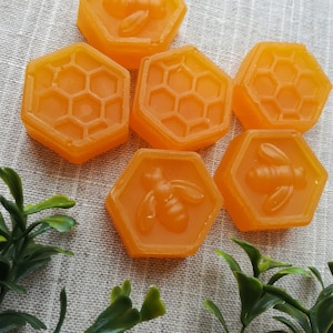Puede incluir: Seis jabones hexagonales con temática de miel con un diseño de abeja y panal. Los jabones son de color naranja claro y están dispuestos en semicírculo sobre una superficie beige texturizada.