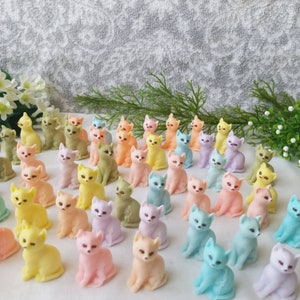 Op de afbeelding: Een verzameling kleine, pastelkleurige kattenbeeldjes in verschillende tinten geel, roze, groen, oranje en blauw. De beeldjes zijn dicht bij elkaar geplaatst, wat een speelse en grillige weergave creëert. De achtergrond is zacht, wit en getextureerd.