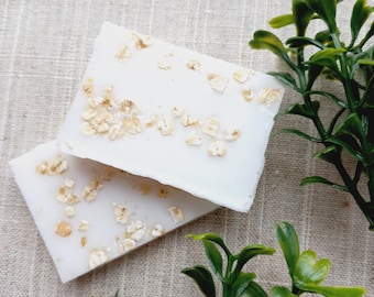 May include: Two white bars of soap with oatmeal flakes on a beige fabric background. Green leafy plants are on either side of the soap.