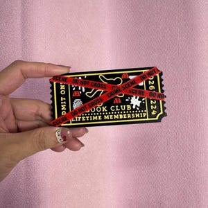 May include: A black and yellow ticket with the text "BOOK CLUB LIFETIME MEMBERSHIP" is crossed with red tape that reads "CRIME SCENE DO NOT CROSS". The ticket has a fingerprint and other crime scene elements.