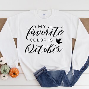 Pode incluir: Camisola branca de manga comprida com o texto "Minha cor favorita é outubro" em fonte cursiva preta com um pequeno gráfico de folha preta.