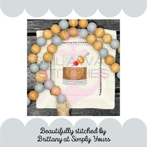 May include: A white linen napkin with a gray and brown beaded garland around it. The napkin has an embroidered image of a cocktail glass with a cherry and a lemon twist. The text "Sail Away Stitches" is embroidered on the napkin.