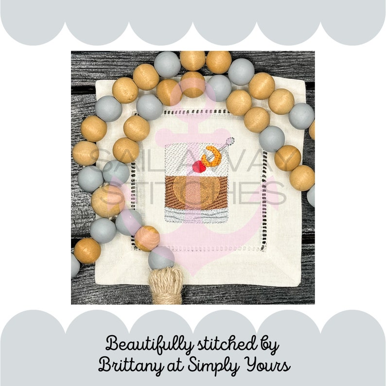 May include: A white linen napkin with a gray and brown beaded garland around it. The napkin has an embroidered image of a cocktail glass with a cherry and a lemon twist. The text "Sail Away Stitches" is embroidered on the napkin.