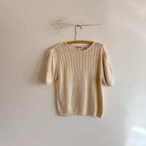 May include: A cream-colored short-sleeved sweater with a cable knit design on the upper chest. The sweater is hanging on a wooden hanger against a plain white wall. The sleeves are slightly puffed.