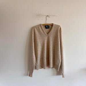 May include: A beige V-neck sweater hangs on a wooden hanger. The sweater has long sleeves and a ribbed hem and cuffs. The fabric appears soft and slightly textured. The sweater is a neutral colour, suitable for various outfits.