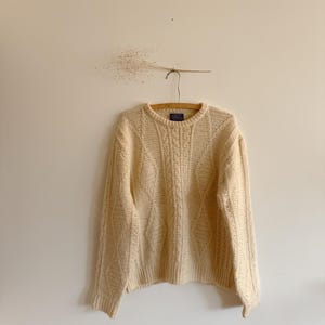 Puede incluir: Suéter de punto trenzado color crema colgado de una percha de madera. El suéter tiene cuello redondo y un patrón texturizado. Se exhibe contra una pared lisa de color blanquecino, creando una estética minimalista. El suéter parece estar hecho de un material suave y cálido.