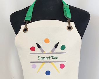 May include: White artist apron with green adjustable straps and a design of two paintbrushes crossed over a palette of colorful paint dots. The text "Sanat Tan" is embroidered on the apron.