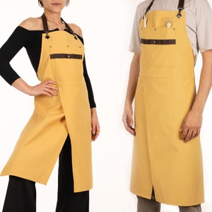 May include: Two yellow canvas aprons with brown leather straps and pockets. The aprons have a split front and are tied at the neck.