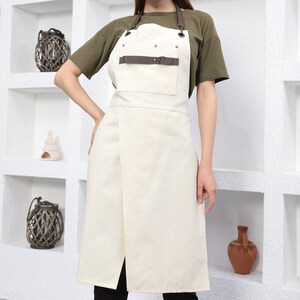 May include: A white canvas apron with brown leather straps and a brown leather pocket. The apron has a long, straight skirt and a single strap that ties around the neck.