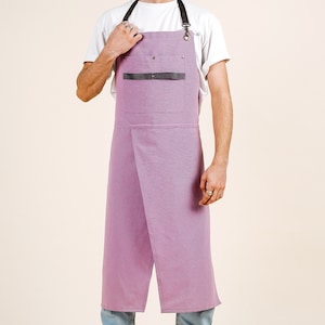 May include: A lavender-coloured apron with a black leather strap and pocket. The apron has adjustable straps and a long, flowing design.