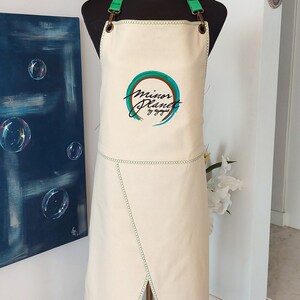 May include: A white canvas apron with green adjustable straps and a green and blue embroidered logo that says "minor planet by ayaje".