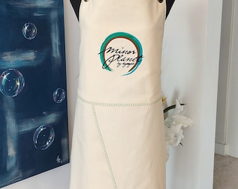 May include: A white canvas apron with green adjustable straps and a green and blue embroidered logo that says "minor planet by ayaje".