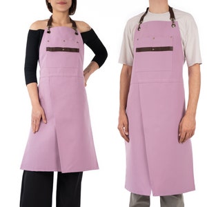 May include: Two pink aprons with brown leather straps and pockets. The aprons have adjustable neck straps and tie closures.