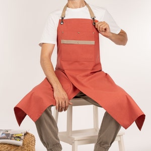 May include: A terracotta-colored apron with a tan pocket and adjustable straps. The apron is made of a durable fabric and has a long, flowing design.