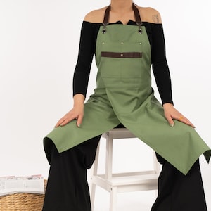 May include: A person wearing a green apron with brown leather straps and a large pocket. The apron is tied around the waist and has a criss-cross strap design in the back.