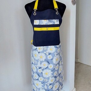 Puede incluir: Un delantal azul marino con tirantes amarillos y estampado de margaritas. El delantal tiene una franja amarilla y un bolsillo en la parte delantera.