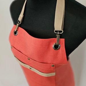 May include: A coral-coloured canvas apron with adjustable straps and a large pocket. The straps are made of a beige canvas material and have metal buckles. The apron has metal rivets and eyelets.