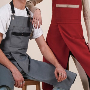 May include: Two people wearing different styles of aprons. One apron is grey with black accents and the other is red with a tan pocket.