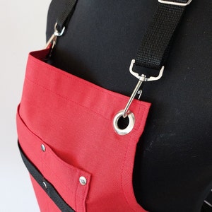 May include: Red canvas apron with adjustable black straps and a large pocket. The apron has a metal ring and a black strap across the front.