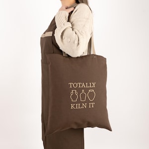 May include: A brown tote bag with a white design that says "Totally Kiln It" with three stylised pottery designs.