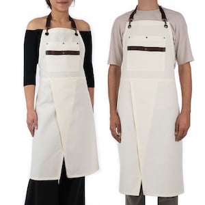 May include: Two white aprons with brown leather straps and a brown leather pocket. The aprons have adjustable straps and a criss-cross back design.