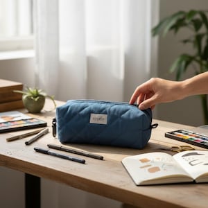 Peut inclure: Une trousse à crayons bleue matelassée avec une fermeture éclair et le nom de la marque "sonation" sur une étiquette blanche. La trousse est sur un bureau en bois avec des stylos, une palette d'aquarelles et un carnet de croquis.