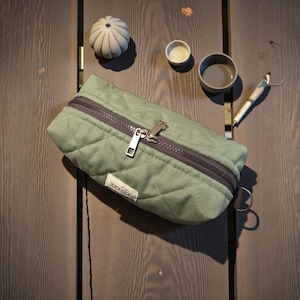 May include: Sage green quilted pencil case with a dark gray zipper and a silver zipper pull. The case has a rectangular shape and a small white label with text. A small decorative pumpkin and two small gray bowls are also visible.